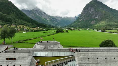 Kids-playing-and-lots-of-tourists-outside-Fjaerland-glacier-museum---Rising-aerial-from-close-to-museum-building-with-exhibition-to-showing-Boyadalen-valley---60-fps-Norway