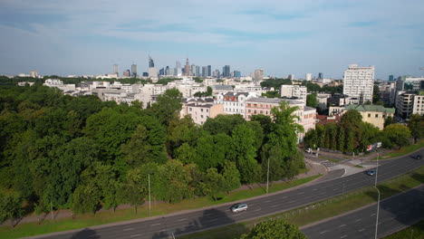 Traffic-through-neighborhood-park-and-Warsaw-city-skyline-in-distance,-aerial