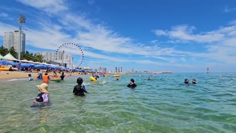 Sokcho-Strand-–-Eine-Menge-Koreanischer-Menschen,-Die-An-Heißen-Sommertagen-Im-Kristallklaren-Meer-Schwimmen,-Riesenrad-Im-Hintergrund-–-POV-Blick-Vom-Wasser-Aus