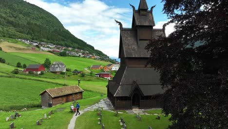 Antena-Exterior-De-La-Iglesia-De-Madera-De-Hopperstad-Durante-El-Verano---Iglesia-Vista-Detrás-De-Un-árbol-Con-Fondo-De-Montaña---Noruega-60-Fps