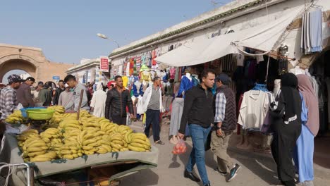 Locals-Walking-Past-Banana-Market-Fruit-Seller-In-Medina,-Essaouira,-On-Sunny-Day