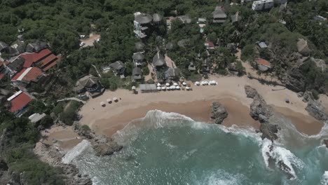 Beach-aerial-tilts-over-grass-roof-resort-hotel-on-rocky-sand-crescent