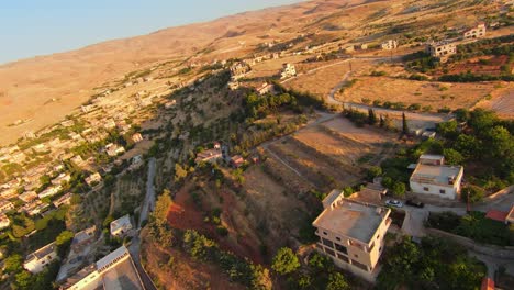 FPV-Drohnenflug-über-Eine-Malerische-Stadt-Auf-Einem-Hügel-Mit-Häusern-Und-Wüstenbergen-Im-Hintergrund-Bei-Sonnenuntergang,-Nahle,-Libanon