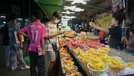 A-group-of-young-male-students-buying-some-snacks-of-mixed-sausages-and-friend-quail-eggs-from-a-street-vendor-at-Chatuchak-Weekend-Night-Market,-in-Bangkok,-Thailand