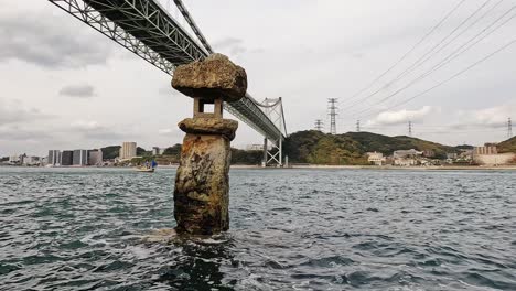 Antigua-Linterna-De-Roca-Japonesa-Medio-Sumergida-En-Agua-Frente-Al-Puente-De-Kanmon-Y-El-Estrecho-De-Kanmon-Entre-La-Isla-Japonesa-De-Honshu-Y-Kyushu