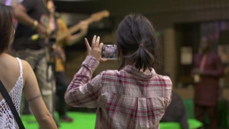 Woman-takes-live-video-capture-and-stream-of-music-concert,-filmed-from-rear-perspective-as-medium-slow-motion-shot