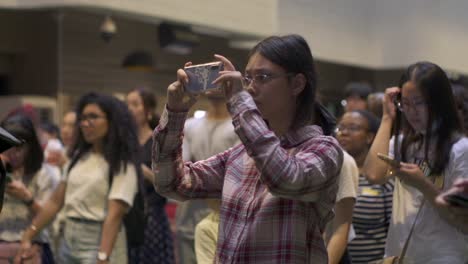 Asian-woman-focused-on-cellphone-video-live-stream-and-recording-during-live-music-performance-with-crowd-in-background,-filmed-as-medium-slow-motion-shot