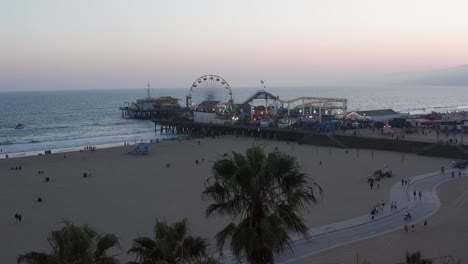 Aerial-close-up-shot-flying-over-palm-trees-to-reveal-the-Santa-Monica-Pier-on-a-bustling-summer-night-in-Los-Angeles,-California-at-twilight