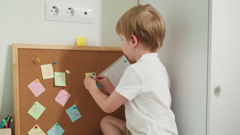 Little-boy-pins-note-to-cork-board-with-stickers-sitting-on-table