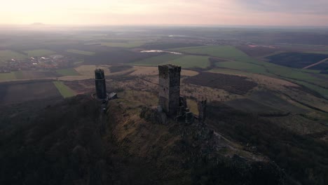 Drone-view-pulling-away,-hilltop-medieval-twin-towers-castle-above-farmlands