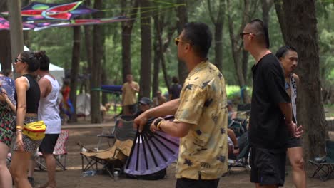 Asian-man-wearing-sun-glasses-and-yellow-shirt-freestyle-dancing-and-waving-Chinese-paper-fan-in-outdoor-ethnic-live-music-festival,-filmed-as-medium-shot-in-slow-motion