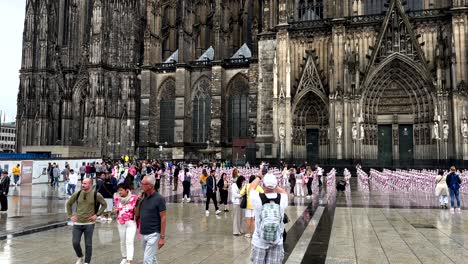 Many-Tourist-visiting-Cologne-Cathedral-with-outdoor-exhibition-artwork-during-rainy-day,-Germany---Slow-motion---child-exploitation-theme-with-pink-dolls
