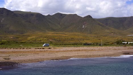 Camper-van-in-green-fields-near-mountain-and-blue-sea-water