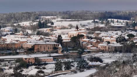 Small-village-under-the-snow-in-France,-Monpazier-in-the-Dordogne,-morning-rises-over-the-countryside,-circular-drone-shot-around-the-church-illuminated-by-the-morning-sun