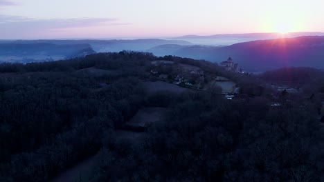 Château-de-Castelnaud-by-drone-at-sunrise-and-in-the-mist,-wide-view-with-the-forest-giving-blue-and-pink-colors-with-violet