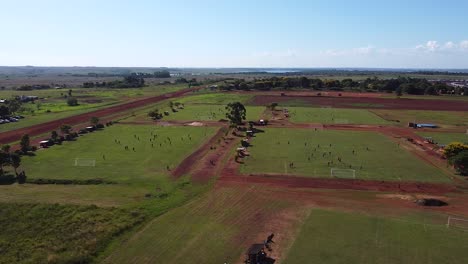 Drone-wide-strafe-past-teams-playing-football-matches-across-multiple-pitches,-sunny-sky-and-landscape-in-background