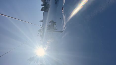 Tilt-up-vertical-handheld-shot-riding-on-a-slow-ski-lift-in-the-Utah-Rocky-Mountains-on-a-bright-sunny-winter-day-with-a-small-train-park-below