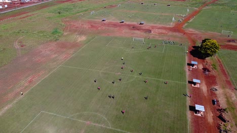 Drone-high-altitude-approach-on-kids-playing-football-match-on-pitch,-goal-kick,-many-other-pitches-in-the-background