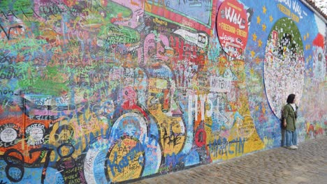 Mujer-Parada-Frente-Al-Muro-De-John-Lennon-En-Praga,-República-Checa:-Graffiti-Inspirado,-Letras-De-Canciones-De-Los-Beatles-Y-Diseños-Relacionados-Con-Causas-Locales-Y-Globales.