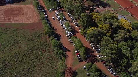 Drone-orbit-over-car-park-in-wooded-area-and-surrounding-fields---cars-drive-past
