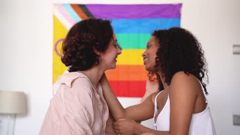 Feliz-Pareja-De-Lesbianas-Diversas-Sentadas-Juntas-Contra-La-Bandera-Del-Arco-Iris