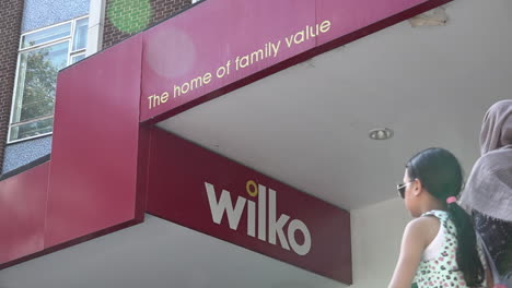 In-slow-motion-people-walk-past-a-Wilko-sign-that-reads,-“The-home-of-family-vale”-on-a-storefront-on-a-busy-high-street-in-Stratford-on-a-hot-summer-afternoon