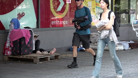 In-slow-motion-people-walk-past-a-barefooted-homeless-person-sitting-with-their-belongings-on-a-wooden-crate-next-to-an-old-mattress-on-a-high-street