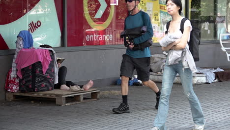 En-Cámara-Lenta,-La-Gente-Pasa-Junto-A-Una-Persona-Sin-Hogar-Descalza-Sentada-Con-Sus-Pertenencias-En-Una-Caja-De-Madera-Junto-A-Un-Colchón-Viejo-En-Una-Calle-Principal