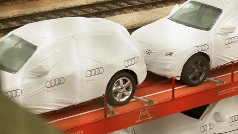 Handkameraaufnahme-Eines-Orangefarbenen-Waggons-Mit-Dem-Audi-Logo,-Der-Zwei-Audi-Wagen-Trägt,-Eingehüllt-In-Eine-Weiße-Schutzhülle,-Vor-Dem-Hintergrund-Von-Bahngleisen-Und-Anderen-Waggons