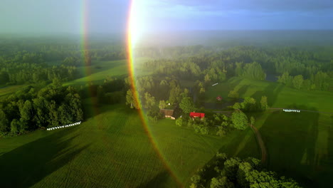 Early-morning-and-rainbow-lens-flare-over-small-countryside-homestead,-aerial-view