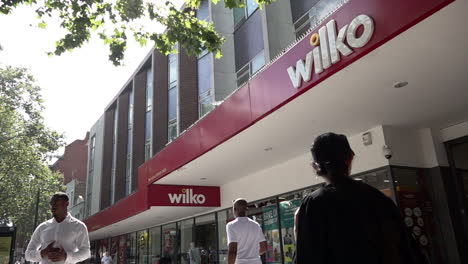 In-slow-motion-people-walk-past-the-storefront-of-a-Wilko-superstore-on-a-busy-high-street-in-Stratford-on-a-hot-summer-afternoon