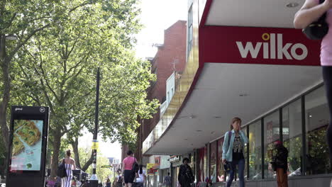People-walk-past-a-Wilko-outlet-storefront-on-a-busy-high-street-in-Stratford-on-a-hot-summer-afternoon