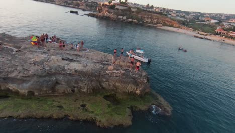 Aerial-orbiting-showing-group-people-on-rocky-island-jumping-into-water-during-sunset-time---FPV-circling-flight