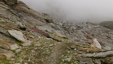 Aerial-backward-view-of-hiker-walking-in-rugged-rocky-mountain-landscape-shrouded-in-fog-in-Valmalenco,-Italy