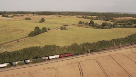Aerial-view-capturing-a-red-locomotive-pulling-white-cargo-cars,-cleaving-through-a-grassed-field-bordered-by-trees-and-a-dirt-road,-amidst-rolling-hills-and-sparse-buildings-under-gray-skies