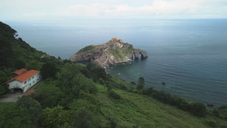 Drone-descends-along-lush-green-sea-cliffs-to-monastery-on-Gaztelugatxe-Basque-Spain,-establishing