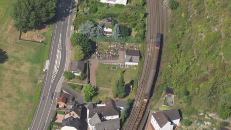 Drone-shot-offering-a-panoramic-view-of-a-quaint-small-town,-interwoven-with-a-bustling-railway-line-and-road,-accentuated-by-charming-white-buildings-with-red-roofs,-surrounded-by-lush-greenery