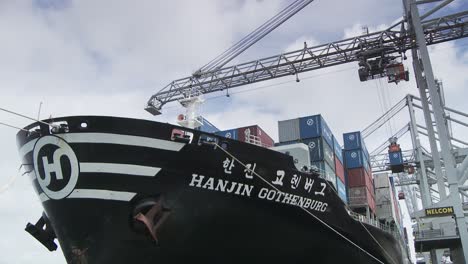 Docked-black-cargo-ship-flaunting,-brimming-with-blue-and-red-shipping-containers,-amidst-an-industrial-setting-under-a-cloudy-sky-with-cranes-above