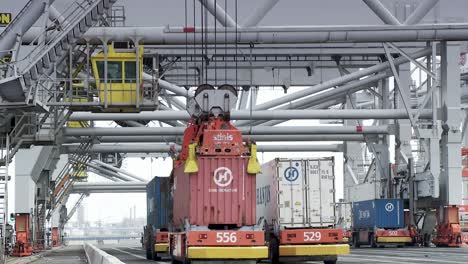 Yellow-crane-lifting-a-red-container-over-multicolored-stacks,-amidst-a-port-setting-with-a-gray-sky,-more-cranes,-shipping-paraphernalia,-and-tire-marked-concrete-foreground