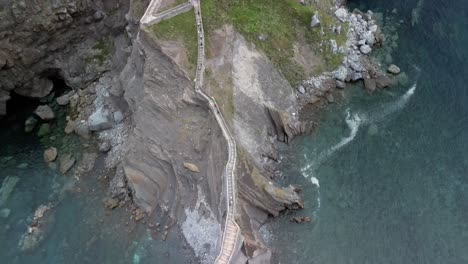 Aerial-descend-pan-up-to-monastery-stairway-on-Gaztelugatxe-Basque-Spain,-cloudy-day-at-sea