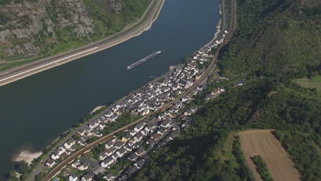 Drone-shot-capturing-a-small-town-with-white-buildings-and-grey-roofs-nestled-by-a-wide-river-with-a-barge,-surrounded-by-green-hills-and-forests,-bisected-by-a-highway