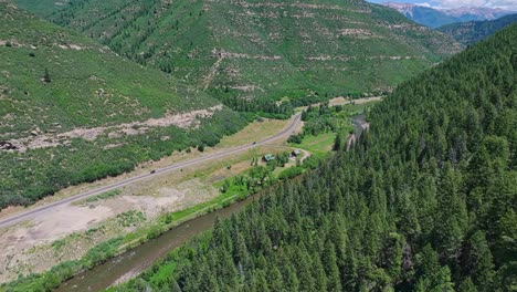 Flying-along-the-Slate-River-and-a-forested-valley-with-a-road-through-the-middle-near-the,-Colorado,-USA