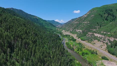 Flying-along-the-Slate-River-and-a-forested-valley-with-a-road-through-the-middle-near-the,-Colorado,-USA