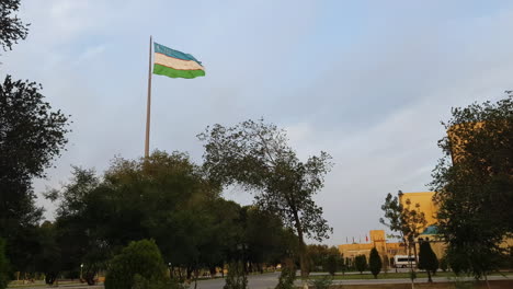 Bandera-Nacional-De-Uzbekistán-Ondeando-En-Un-Poste-En-El-Centro-Del-Parque-De-La-Ciudad,-Vista-Amplia