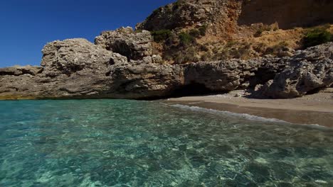 Quiet-beach-sheltered-by-cliffs-on-shore-of-Ionian-sea,-clean-emerald-seawater-reflecting-summer-sunlight