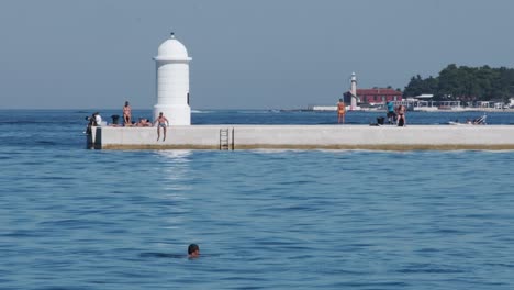 Gente-Tomando-El-Sol-En-El-Paseo-Marítimo-De-Zadar-Con-Faro-Y-Faro-Lejano,-Verano-En-Croacia