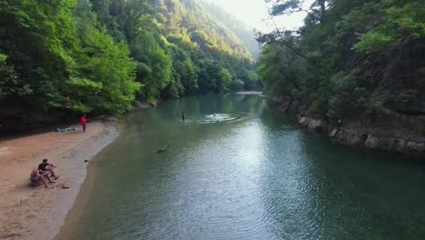 Beautiful-woman-in-sexy-swimsuit-smiles-and-waves-as-drone-takes-off-soaring-above-river-in-canyon