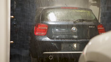 Handheld-camera-captures-a-black-BMW-car,-awash-with-soapy-suds-and-water-in-a-car-wash,-surrounded-by-swirling-brushes-and-water-jets