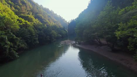 Drone-fly-through-of-river-at-base-of-canyon-in-shadow-of-ridgeline-trees,-sun-bursts-above