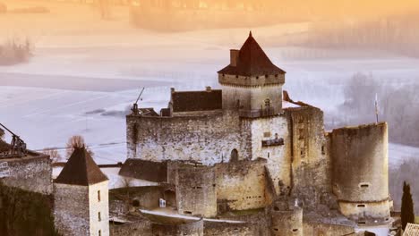 Luftaufnahme-Des-Schlosses-Castelnaud-Am-Morgen,-Nahaufnahme-Der-Architektur-Dieses-Prächtigen-Schlosses-Am-Flussufer-In-Orangefarbenem-Und-Blauem-Licht